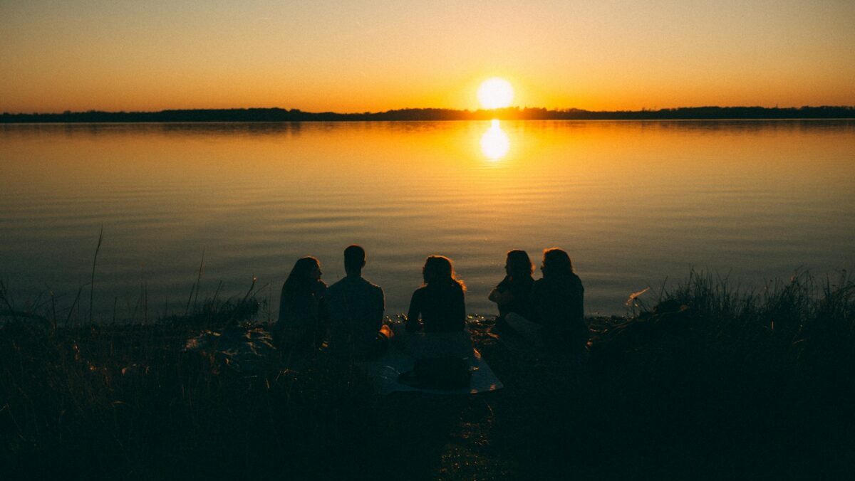 a group of people sitting on the shore of a lake at sunset